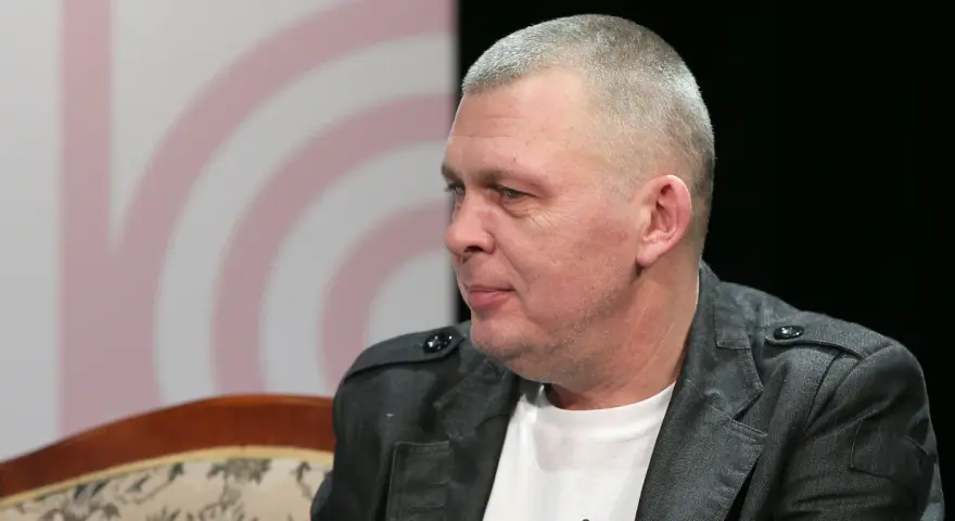 Adam z zespołu Raz Dwa Trzy w szarej marynarce, siedzi na kanapie.