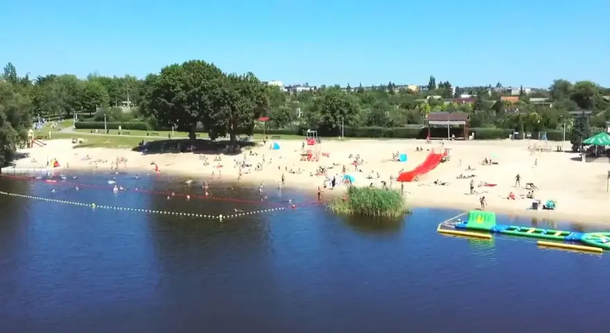 Słoneczny dzień nad jeziorem Grzymisławskim. Ludzie odpoczywają na plaży, kąpią się i bawią na dmuchanych atrakcjach.