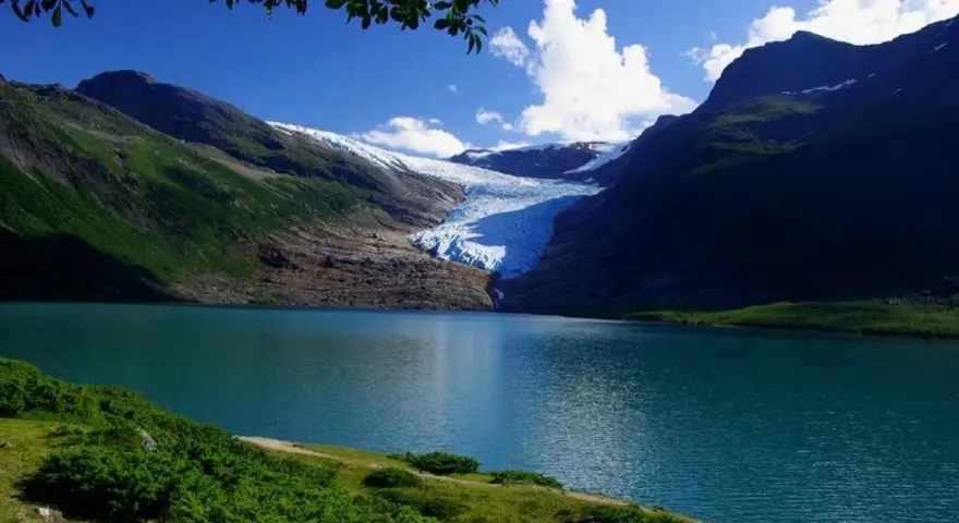 Blå sjö vid foten av en glaciär i Steinkjer, Norge. Grönskande bergssluttningar möter det turkosa vattnet under en klarblå himmel.