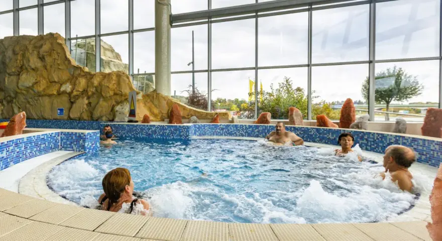 Ludzie relaksują się w basenie termalnym z hydromasażem, otoczonym skalnymi formacjami i dużymi oknami.