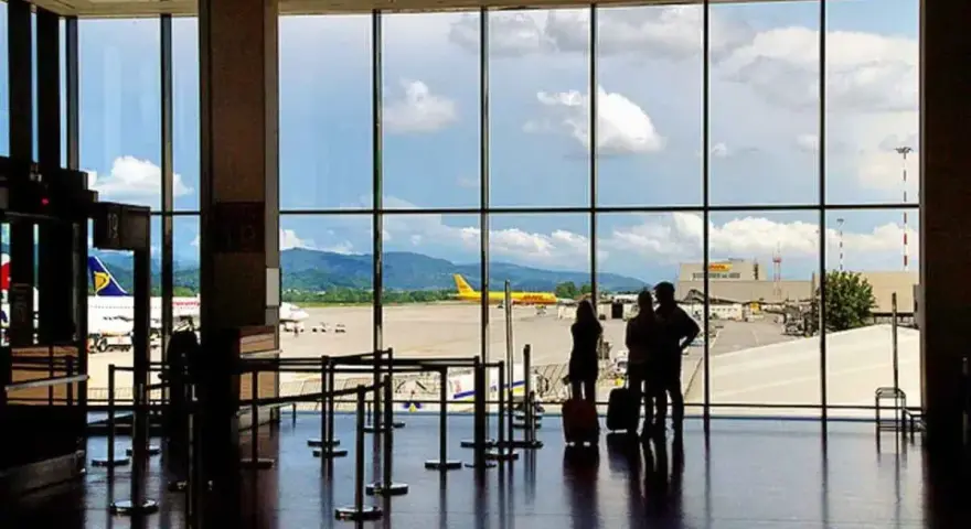 Widok z okna na lotnisko Bergamo. Trzy osoby z walizkami czekają na samolot. W tle żółty samolot DHL i góry.
