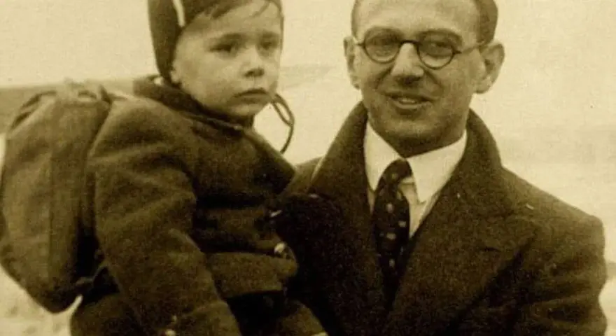 Nick Winton porte un enfant sur son bras, un petit garçon avec un sac à dos, prêt pour un nouveau départ.