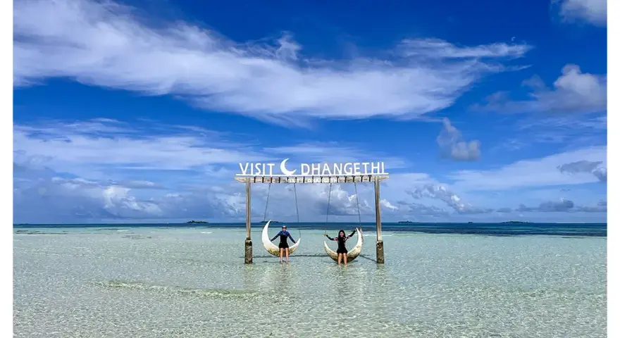 Dos personas disfrutan de columpios en forma de luna sobre aguas cristalinas. Un cartel dice "Visit Dhangethi", mostrando que las Maldivas low cost son posibles.