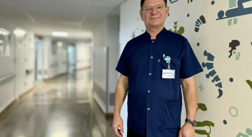 Mężczyzna w granatowym uniformie medycznym stoi na tle szpitalnego korytarza z kolorowymi grafikami.