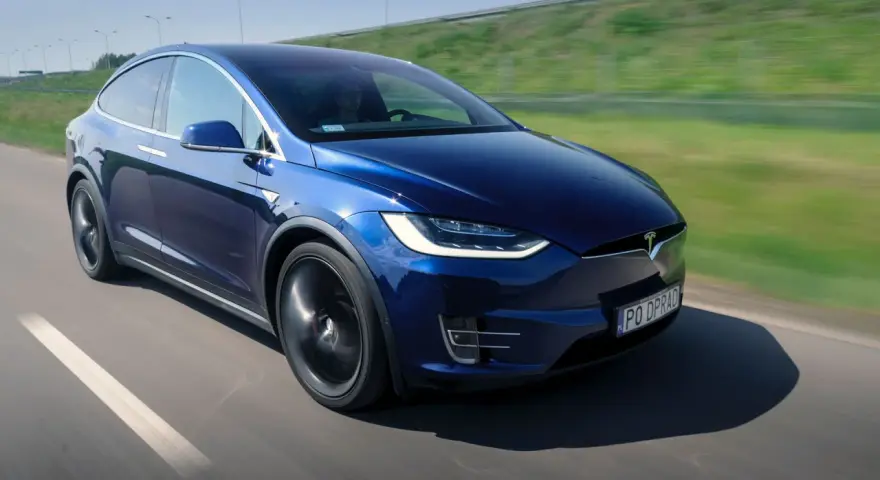 Granatowy Tesla Model X w ruchu na drodze, z widocznym kierowcą.