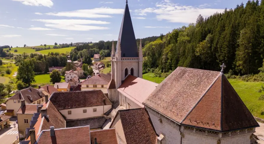 Vue aérienne d'un village pittoresque du haut Doubs, dominé par une église à la flèche élancée, entouré de collines verdoyantes et de forêts.