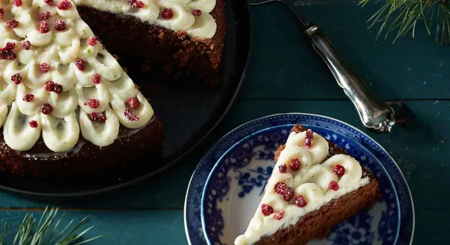 En mjuk pepparkaka med fluffig frosting och röda bär. En bit är upplagd på en blåvit tallrik, redo att avnjutas.