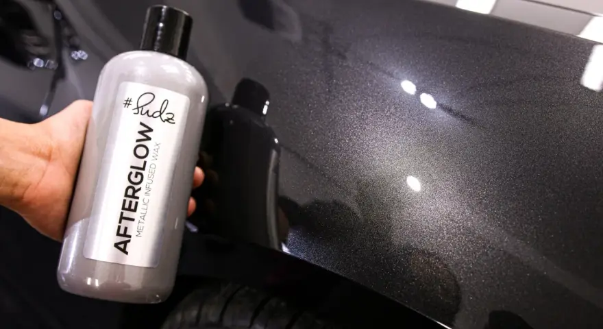 Bottle of "Afterglow Metallic Infused Wax" held against a dark, metallic car finish, showcasing the product's ability to create multiple coats of wax for a deep shine.