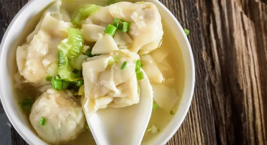 Aromatyczne pierożki wonton w bulionie, ozdobione szczypiorkiem i kawałkami warzyw, na drewnianym tle.