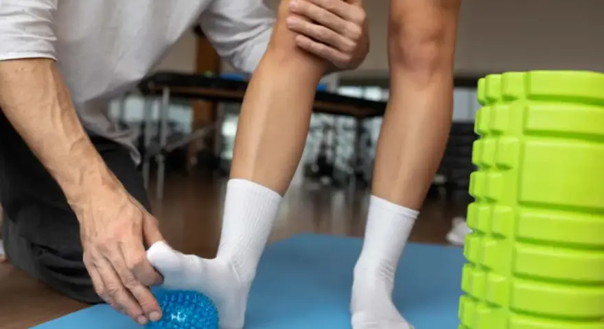 Fizjoterapeuta pomaga w rehabilitacji stawu skokowego, masując stopę pacjenta niebieską piłeczką. Obok stoi zielony wałek do masażu.
