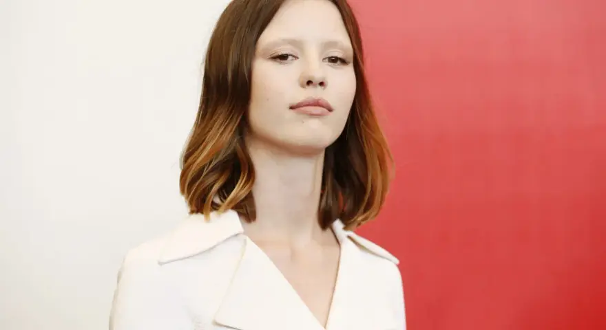 La giovane attrice protagonista del film di mia goth, con capelli castani mossi e un'espressione intensa, indossa un abito bianco.
