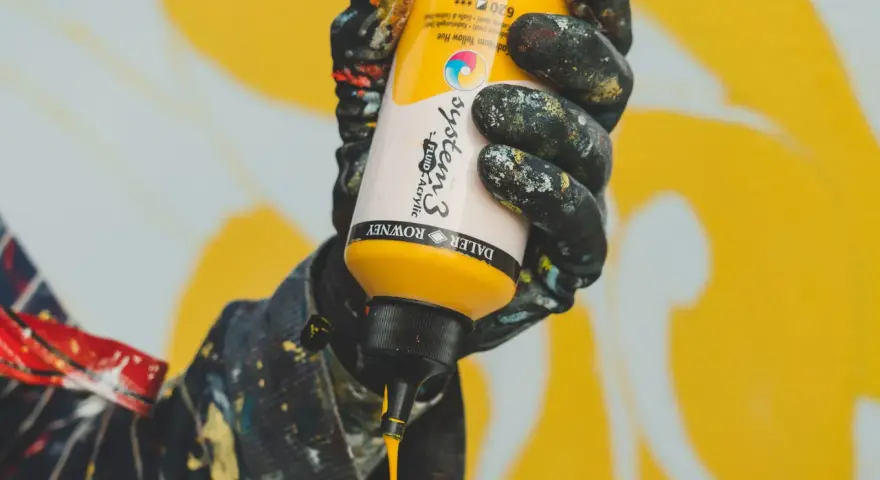 Main jaune de peinture acrylique Daler Rowney System 3, versée par une main gantée, pour un mélange de couleurs.