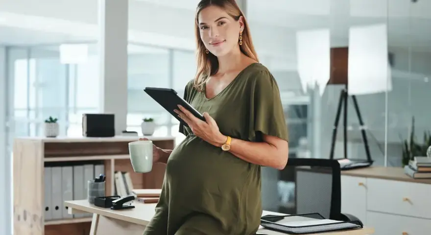 Uśmiechnięta ciężarna kobieta z tabletem i kubkiem w biurze. Dowiedziałam się o ciąży po okresie wypowiedzenia, ale teraz planuję przyszłość.