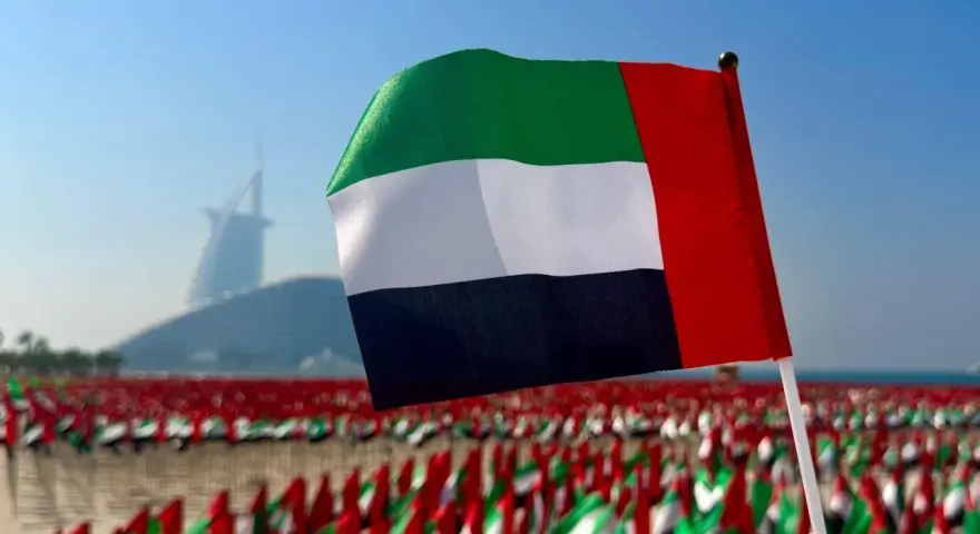 Flaga Zjednoczonych Emiratów Arabskich powiewa na tle drapaczy chmur w Dubaju. W tle widać morze małych flag.
