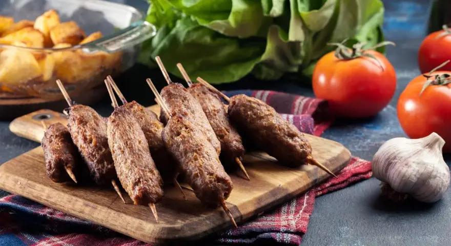 Aksamitne ćevapi na drewnianej desce, obok pomidory, czosnek i pieczone ziemniaki. Pyszny posiłek gotowy do podania.