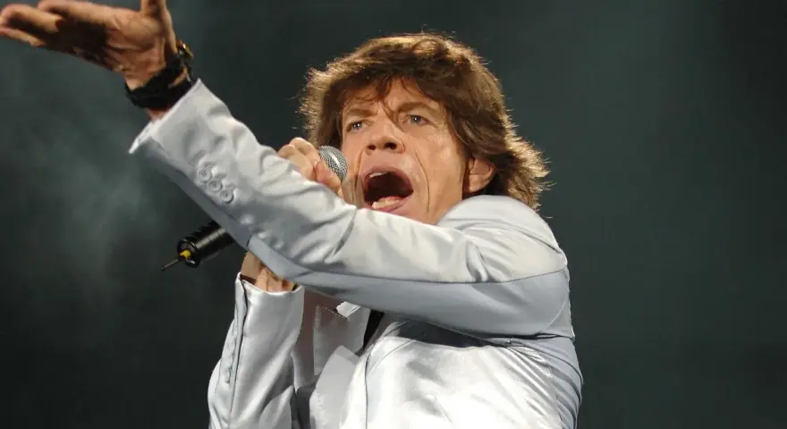 Mick Jagger, wokalista zespołu, śpiewa z mikrofonem, gestykulując ręką.