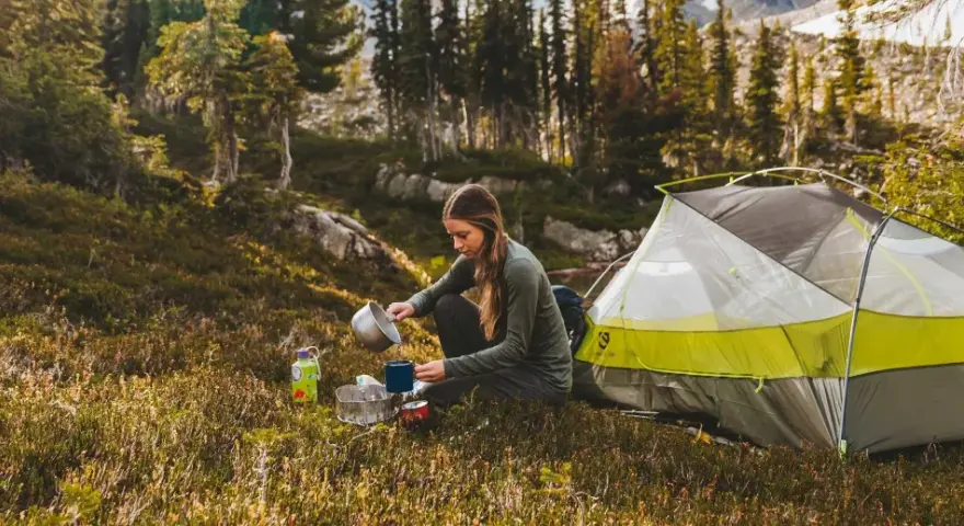 A woman enjoys a peaceful moment of camping alone, preparing a warm drink next to her tent amidst a serene forest landscape.