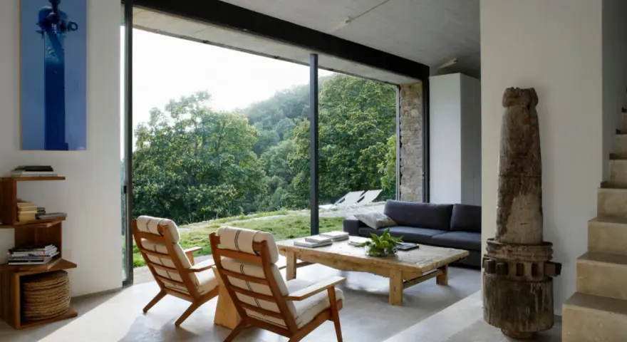 Salón moderno con sofás, sillones y mesa de centro rústica. Los suelos de piedra, con un estilo que evoca los años 70, contrastan con la vista exterior de un bosque frondoso.