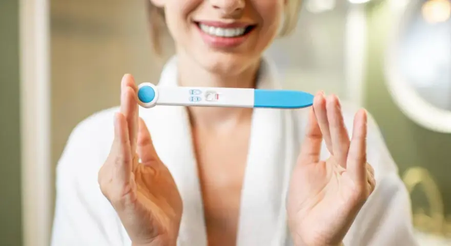 Uśmiechnięta kobieta w szlafroku trzyma test ciążowy z dwoma kreskami. To moment, kiedy warto zastanowić się, kiedy zrobić test ciążowy.