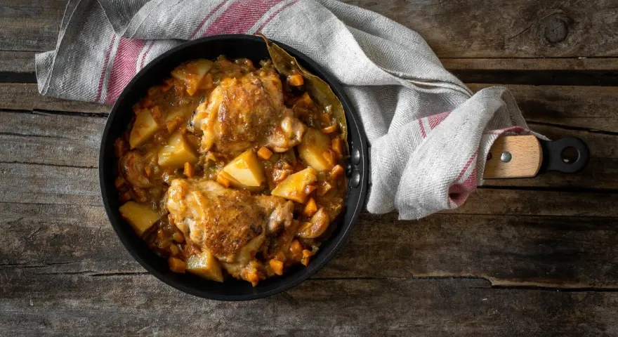 Estofado de pollo con patatas y zanahorias en sartén sobre mesa de madera.