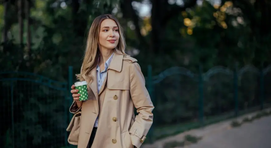 Donna con trench beige, un classico del marchio Burberry, sorseggia un caffè all'aperto.