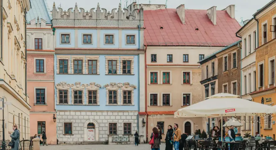Kolorowe kamienice na rynku, idealne na tani city break. Ludzie spacerują, siedzą w ogródku kawiarnianym.