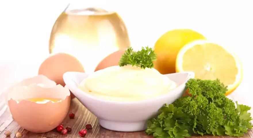 Un bol de mayonnaise maison crémeuse, entouré d'œufs, de citron, d'huile et de persil.