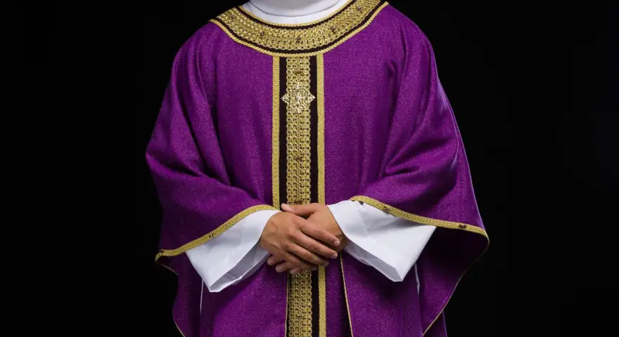 Eleganckie fioletowe szaty liturgiczne z bogatymi złotymi zdobieniami, noszone przez mężczyznę z dłońmi złożonymi na piersi.