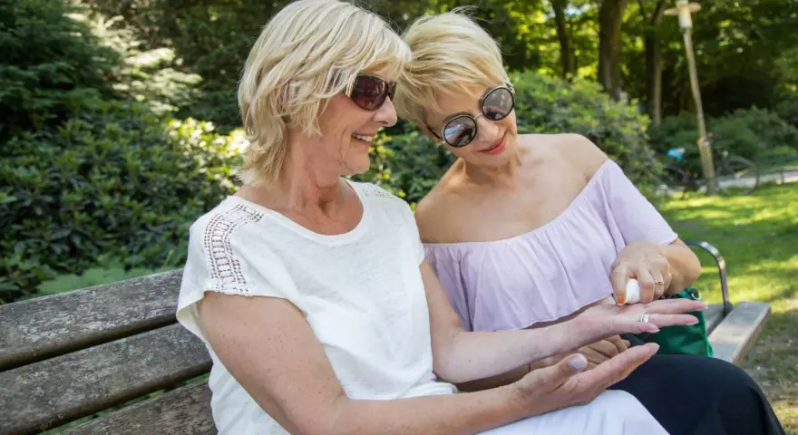 Zwei Frauen, beide über 60, sitzen auf einer Parkbank. Eine trägt eine weiße Bluse, die andere ein lila Off-Shoulder-Oberteil. Die eine Frau trägt Sonnencreme auf die Hand der anderen auf.