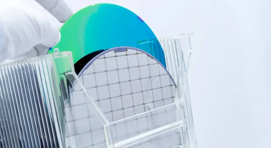 A gloved hand holds a silicon wafer, its grid pattern visible. Another wafer, iridescent blue and green, rests behind it. This image explores the question: is plastic a semiconductor?
