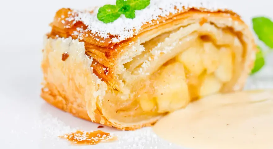 Ein Stück Apfelstrudel mit Filoteig, bestäubt mit Puderzucker und garniert mit Minze, serviert mit Vanillesauce.