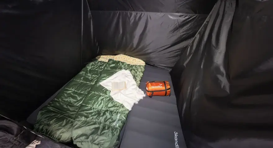 Schlafsack und Kissen auf der Isomatte, welche Seite oben ist, ist klar erkennbar. Ein kleines orangefarbenes Päckchen liegt daneben.