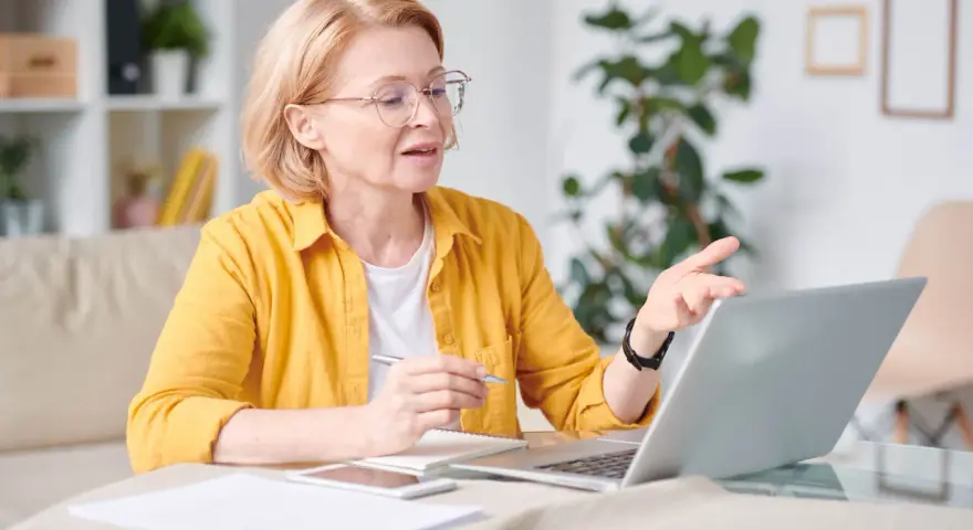 Kobieta w okularach i żółtej koszuli pracuje przy laptopie, trzymając długopis. To jej dodatek funkcyjny do pracy zdalnej.