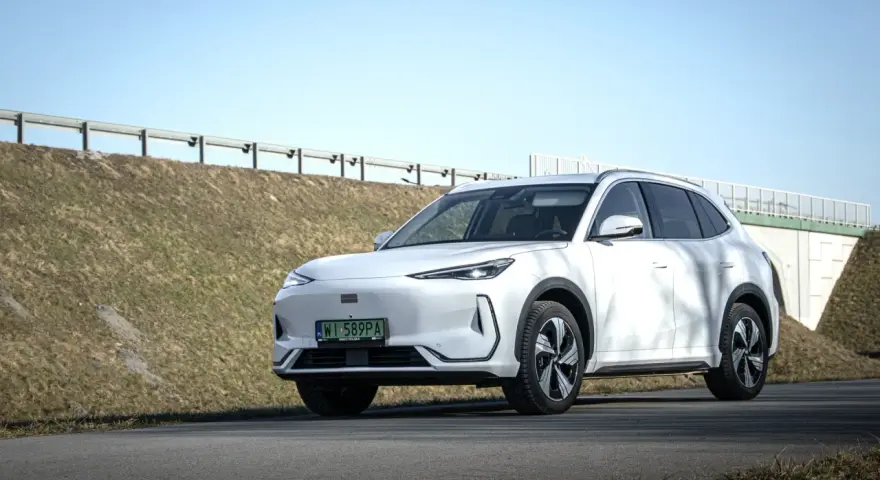 Biały SUV Geely E5 (EX5) na tle wiaduktu i niebieskiego nieba.