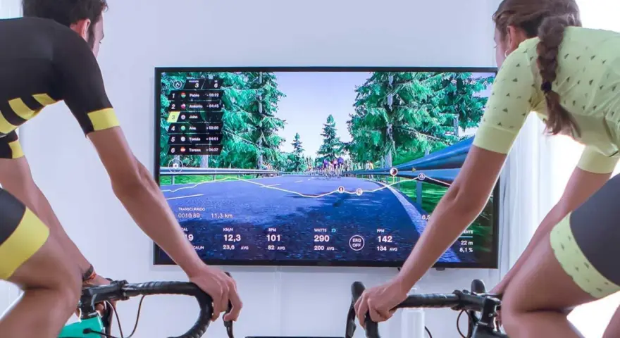 Two cyclists race virtually on a scenic road using indoor cycling apps. The screen displays their stats and rankings.