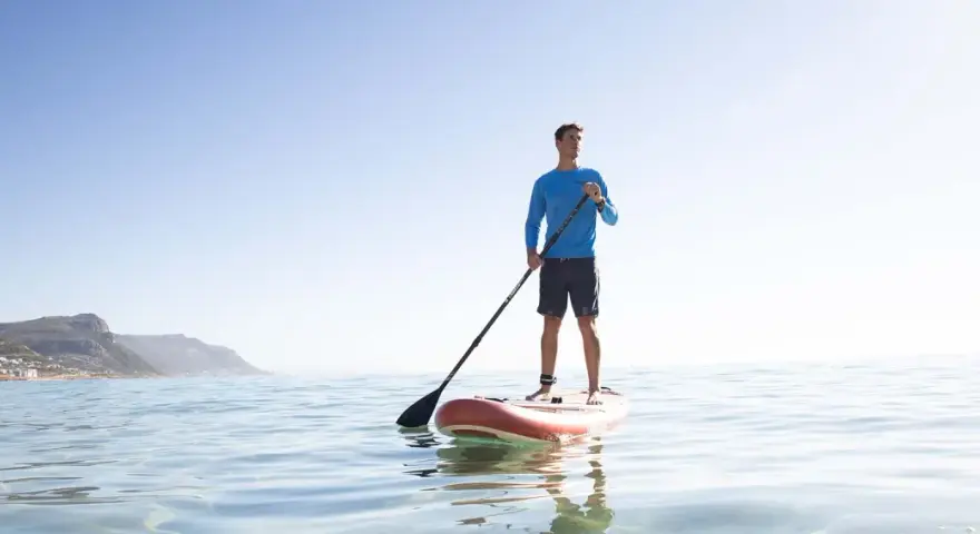 Mann steht auf SUP-Board, hält Paddel richtig. Ruhige See, bergige Küste im Hintergrund.