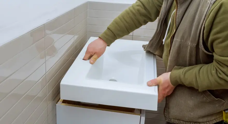Installation d'un lavabo blanc. L'installateur vérifie la hauteur de l'arrivée d'eau du lavabo avant de le fixer.