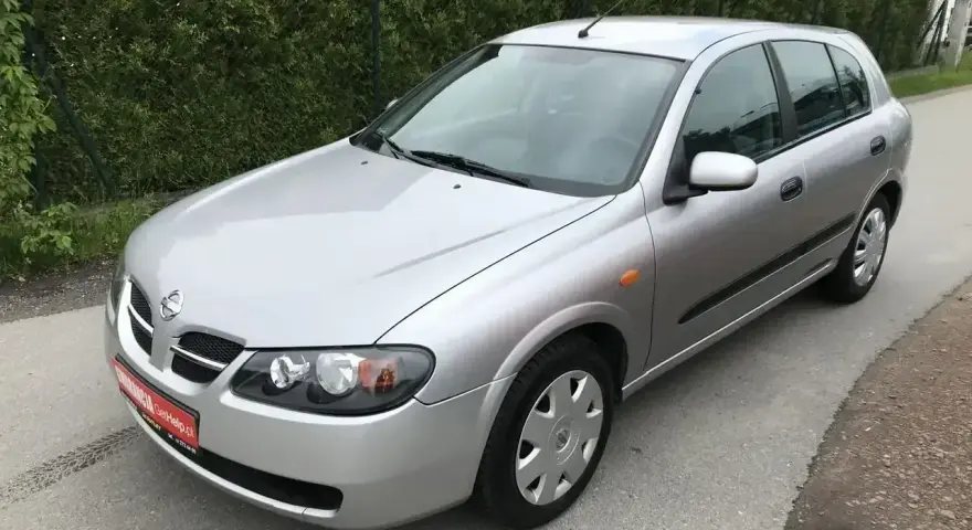 Srebrny Nissan Almera hatchback stoi na asfaltowej drodze, otoczony zielenią.