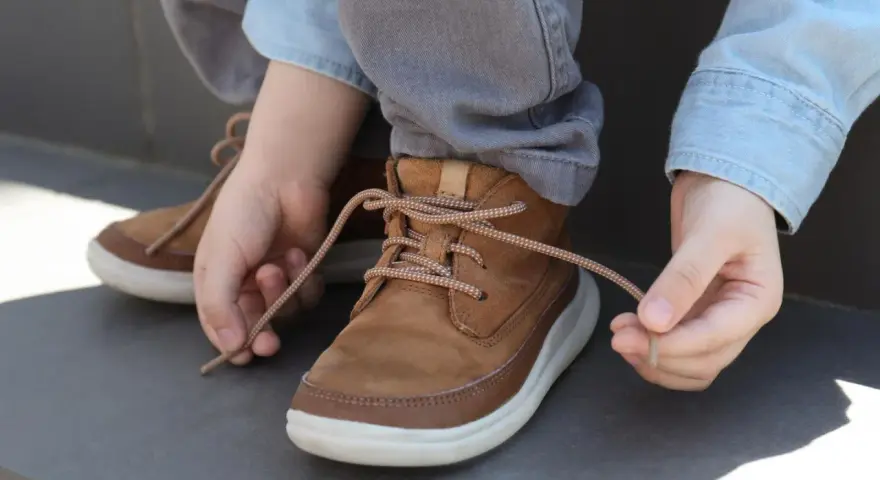 Kind lernt, seine braunen Schnürstiefel zu binden. Die Schnürsenkel sind hellbraun und gemustert.
