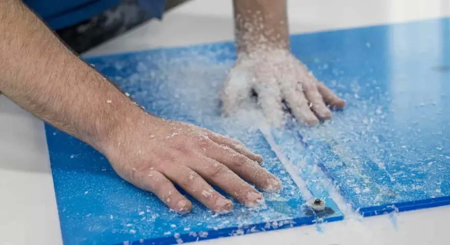 Hands guide a router, creating a clean cut on a blue plexiglass sheet, leaving a trail of white shavings. This is how you cut plexiglass with a router.