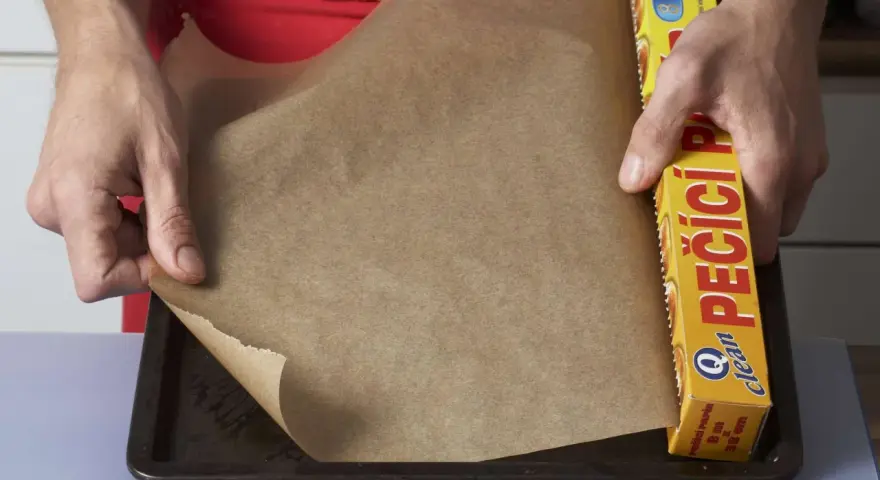 Hände legen Backpapier auf ein Blech. Die Rolle mit der Aufschrift "PEČÍCÍ PAPÍR" zeigt, dass man Backpapier einfetten muss, um ein Anhaften zu verhindern.