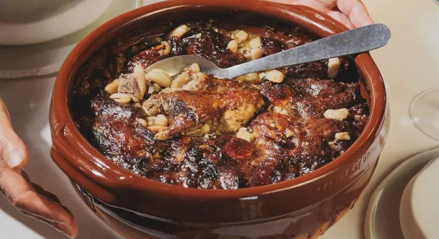 Un plat en terre cuite débordant de haricots blancs, de morceaux de viande dorée et de saucisses, prêt à être dégusté. La recette du vrai cassoulet traditionnel, un délice réconfortant.