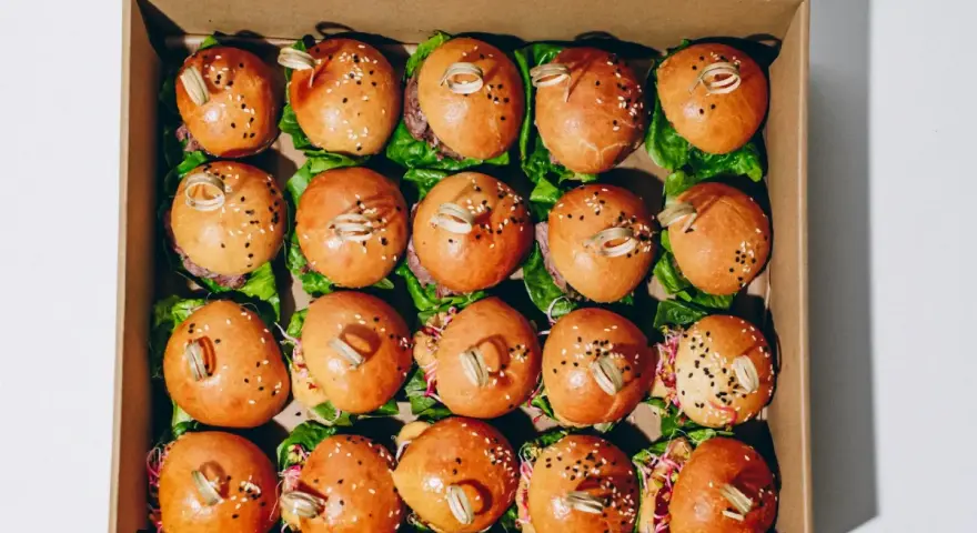Pudełko z dwudziestoma mini burgerami na liściach sałaty, posypane sezamem i ozdobione patyczkami.