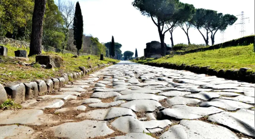 Ancient Roman cobblestone road lined with trees and ruins, leading towards the horizon.