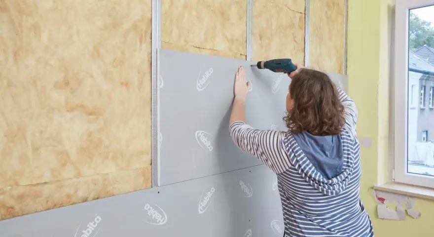 Frau schraubt Rigipsplatten an eine Wand mit Dämmung. Die Platten werden auf Metallschienen befestigt.