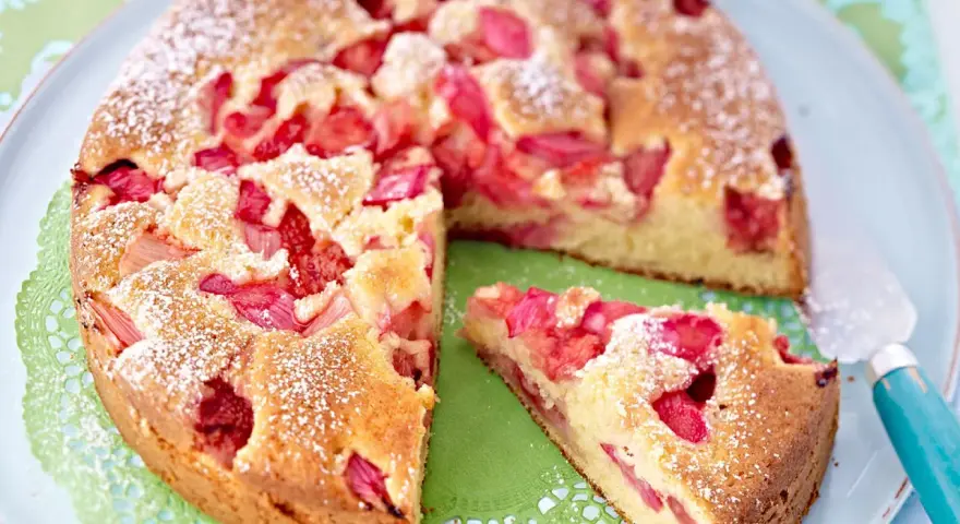Ein saftiger Erdbeer Rhabarber Kuchen, bestäubt mit Puderzucker, wartet auf seinen Anschnitt. Ein Stück ist bereits auf einem grünen Tupfer platziert.