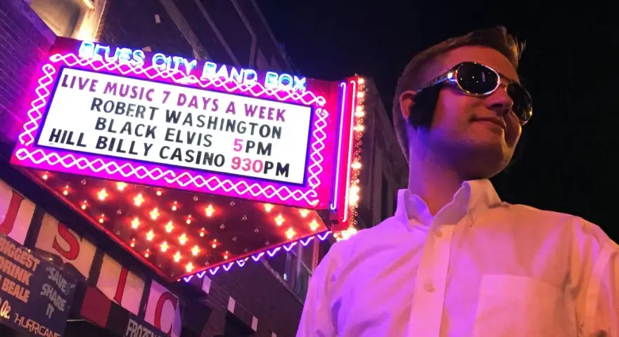 Mann im Elvis-Stil mit Sonnenbrille vor der "Blues City Band Box"-Neonreklame, die "Black Elvis" ankündigt.