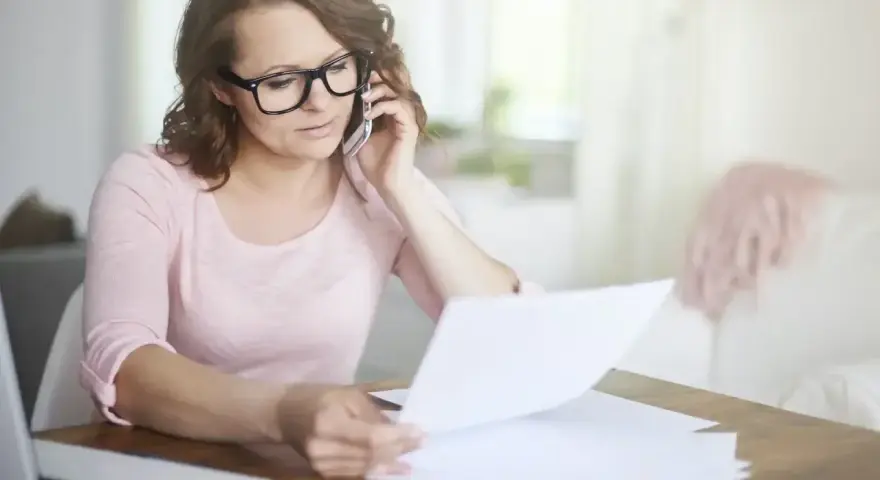 Kobieta w okularach przez telefon wyjaśnia sprostowanie świadectwa pracy, przeglądając dokumenty przy biurku.