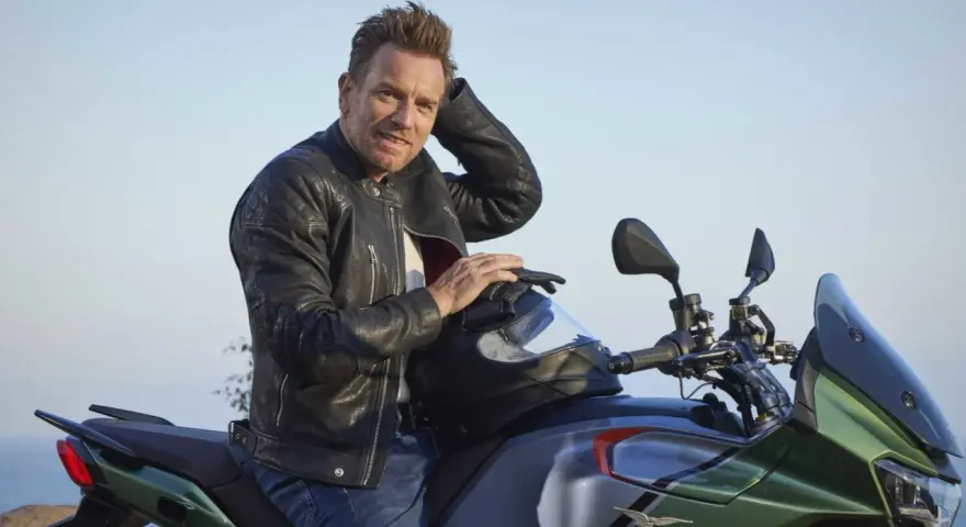 Ewan McGregor, con giacca di pelle, posa su una Moto Guzzi verde e grigia, pronto per un'avventura.