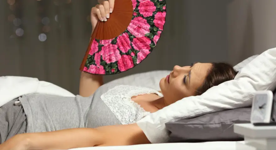 Frau liegt im Bett, nass geschwitzt, fächelt sich mit einem Blumenfächer Luft zu. Die Nacht ist heiß.